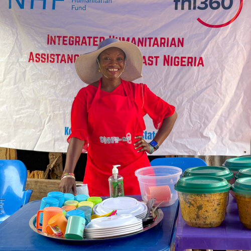A woman in a red apron stands behind tubs of prepared food.