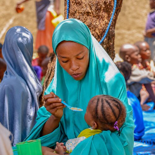 A woman wearing teal spoonfeeds a baby food.